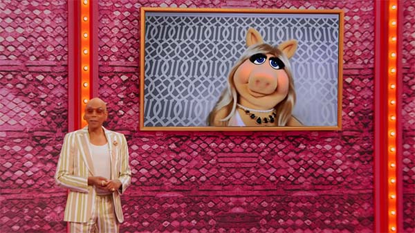 RuPaul on the set of a TV show, with Miss Piggy appearing on a large monitor on the wall