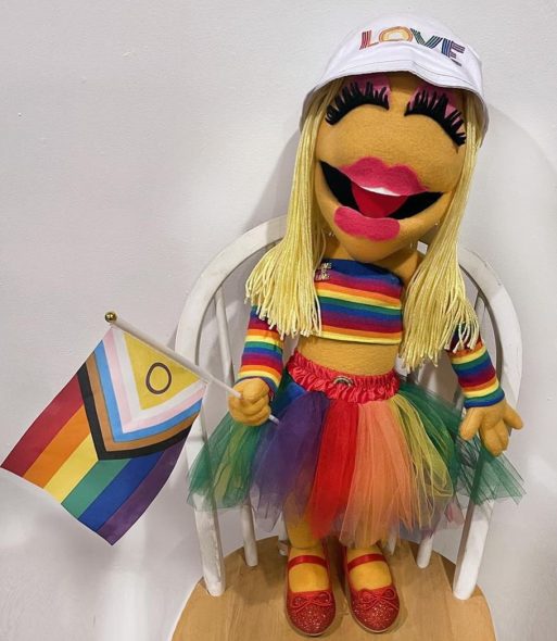 A handmade plush replica of Janice wearing rainbow-colored clothing, a hat reading LOVE, and sparkly red shoes; and holding an Inclusive Pride Flag