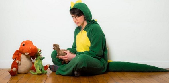 Kira Hall sits on the floor, wearing a dinosaur costume with toy dinosaurs nearby