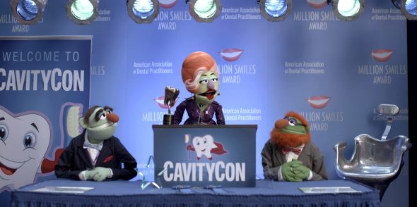 Dr. Tina Teeth stands behind a lectern, speaking at CavityCon, with her husband and her son (Dr. Teeth) sitting on either side of her.