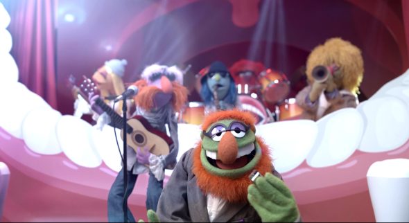Dr. Teeth stands in front of the rest of the Electric Mayhem, holding a gold floss box