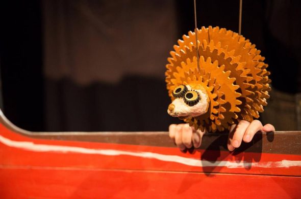 A hedgehog puppet made of cardboard and various other materials suspended over a puppet stage above two human hands.