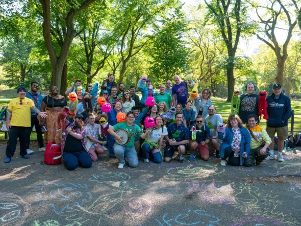 Several Muppet fans gathered at the Jim Henson bench in Central Park. Many of them are holding puppets, and there are chalk drawings of Muppets all over the ground.